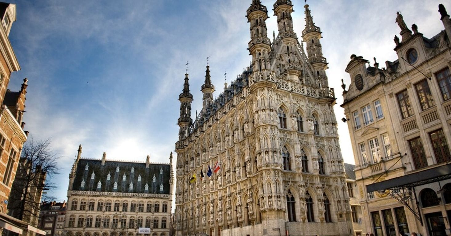 A historic ornate Gothic building with multiple spires stands next to a similarly intricate structure under a blue sky with scattered clouds. Flags are displayed on the building's facade.