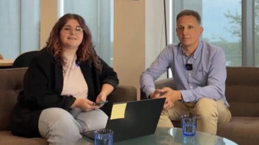 Two people are seated on a couch in an office setting with a laptop and water glasses on the table in front of them.