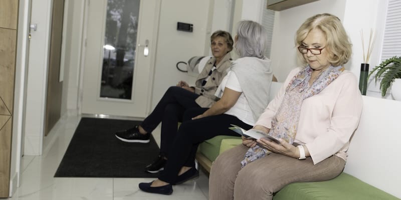 Three women sitting in a waiting area; two on one side, one on the other reading a booklet. The room has a door, mat, and plant decor.