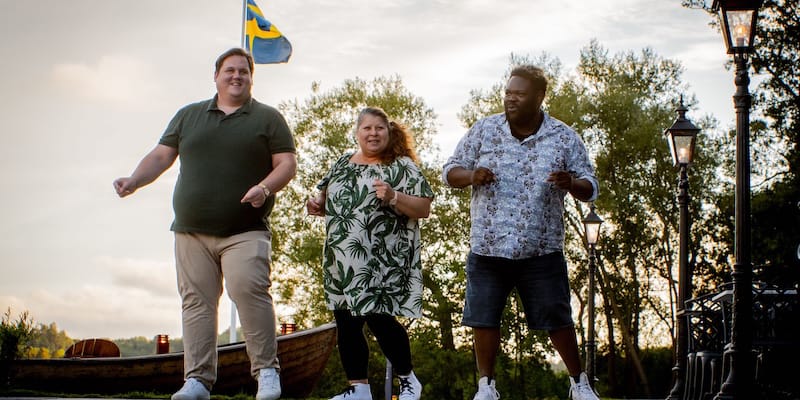 Three people standing outdoors, smiling, with trees and a Swedish flag in the background.