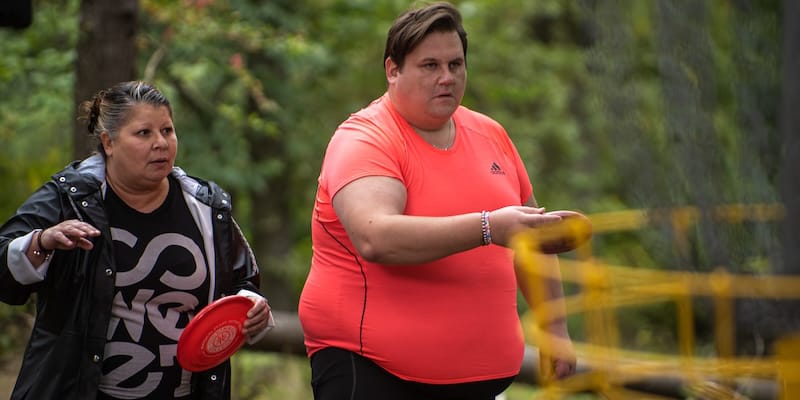 Two people in casual clothing holding frisbees, appear focused during a game in an outdoor setting with trees and a disc golf basket visible.