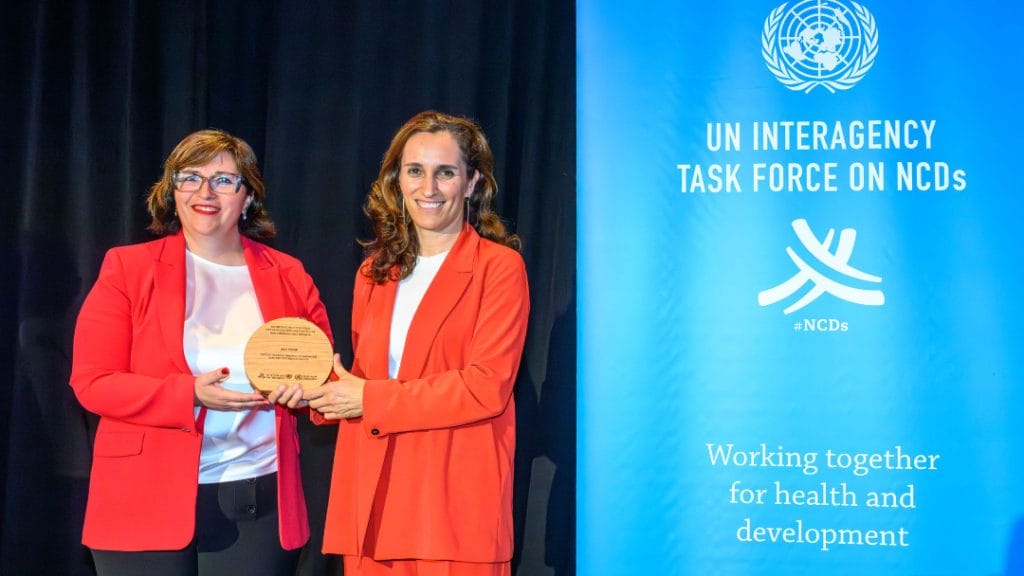 Two women in red blazers hold a round award next to a blue UN Interagency Task Force on NCDs banner.