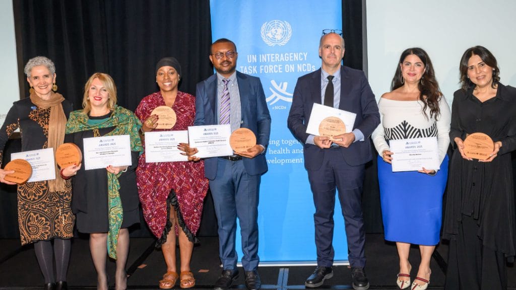 Seven people stand in a row holding certificates and round plaques in front of a UN Interagency Task Force on NCDs banner.
