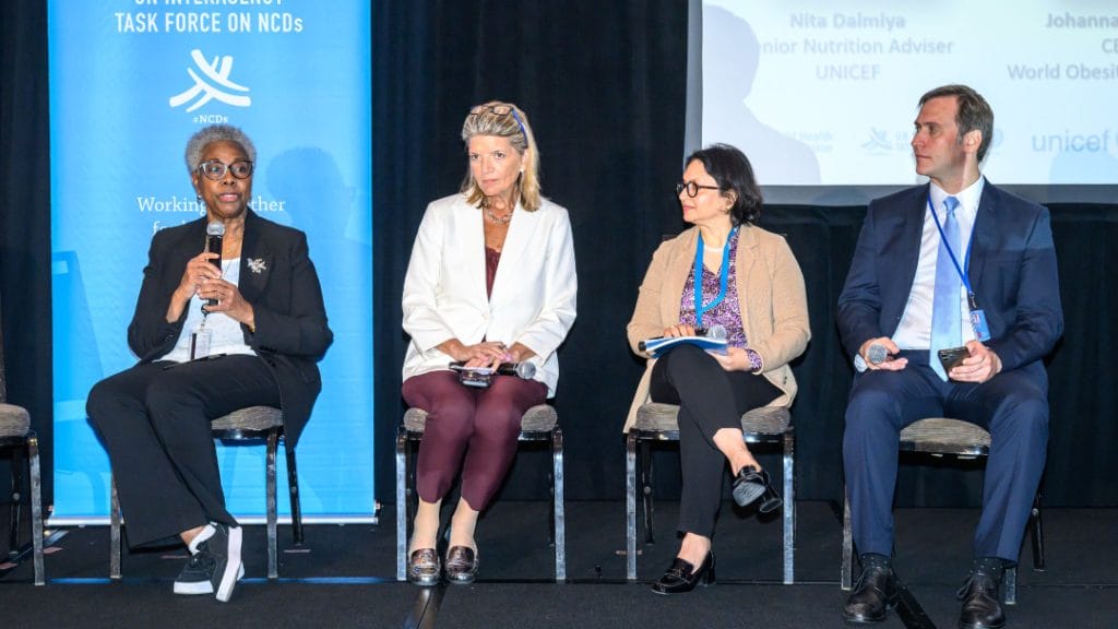 Four panelists sit on stage at a conference, listening and holding microphones or notepads, with a Task Force on NCDs banner in the background.