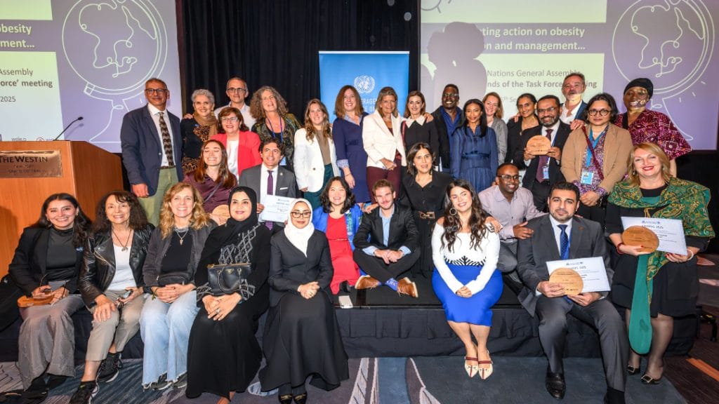 A large group of men and women pose indoors at an event, with some holding plaques and certificates; presentation slides are displayed in the background.