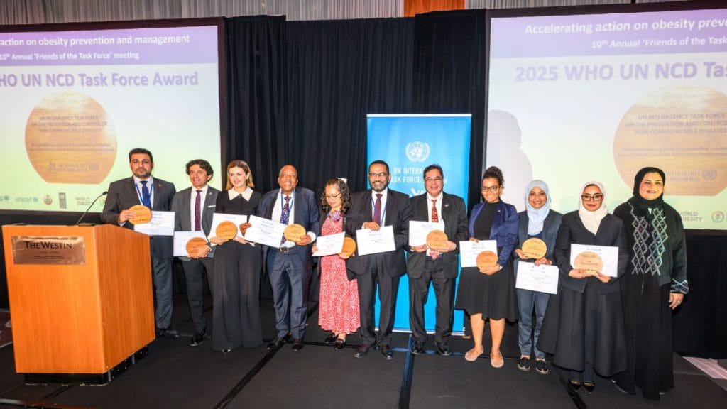 A group of eleven people stands on stage holding plaques and certificates in front of a screen displaying "2025 WHO UN NCD Task Force Award.