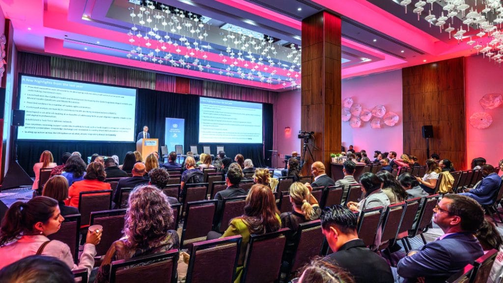 A large audience sits facing a stage where a speaker presents slides in a modern, well-lit conference room with pink and purple lighting.