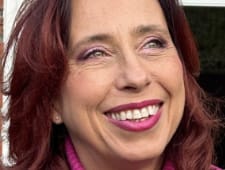 A woman with reddish-brown hair, wearing bright pink lipstick and eye makeup, smiles while looking slightly upwards. She is dressed in a pink top.